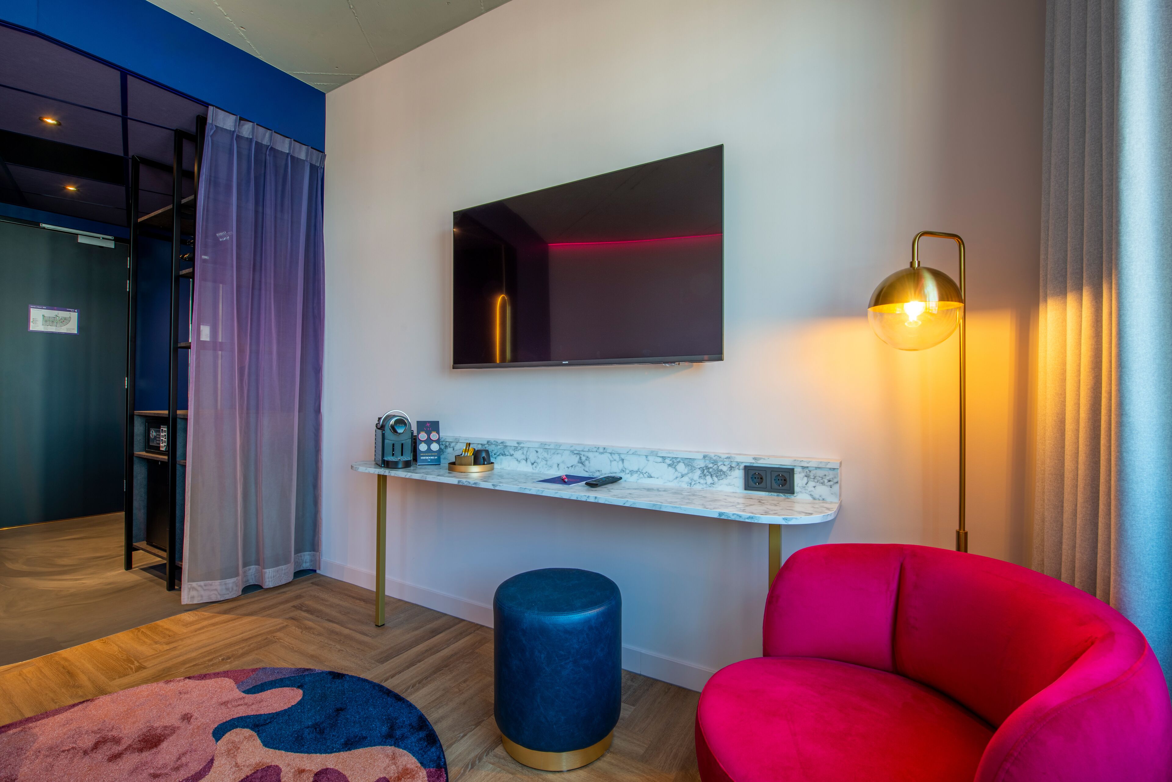 Ein modernes Hotelzimmer mit Flachbildfernseher, Schreibtisch mit Marmorplatte, rotem Sessel, blauem Hocker und Stehlampe.