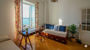 A room with a wooden desk, a bed with blue and white bedding, a potted plant, and a view of the sea through a window with floral curtains. - Casa Baia Salata 1 (Imperia)
