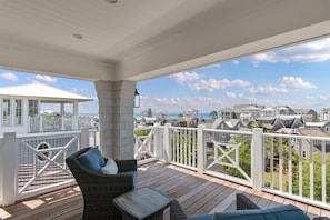 A covered outdoor deck with wicker furniture, a view of a coastal community, and a white railing.