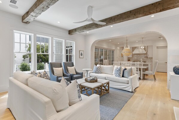 A spacious living room with a white sofa, armchairs, and a coffee table. There are large windows with white shutters, a ceiling fan, and a kitchen area visible in the background.