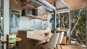 Een moderne keuken met een centraal eiland, houten vloeren en uitzicht op het groen door grote ramen.