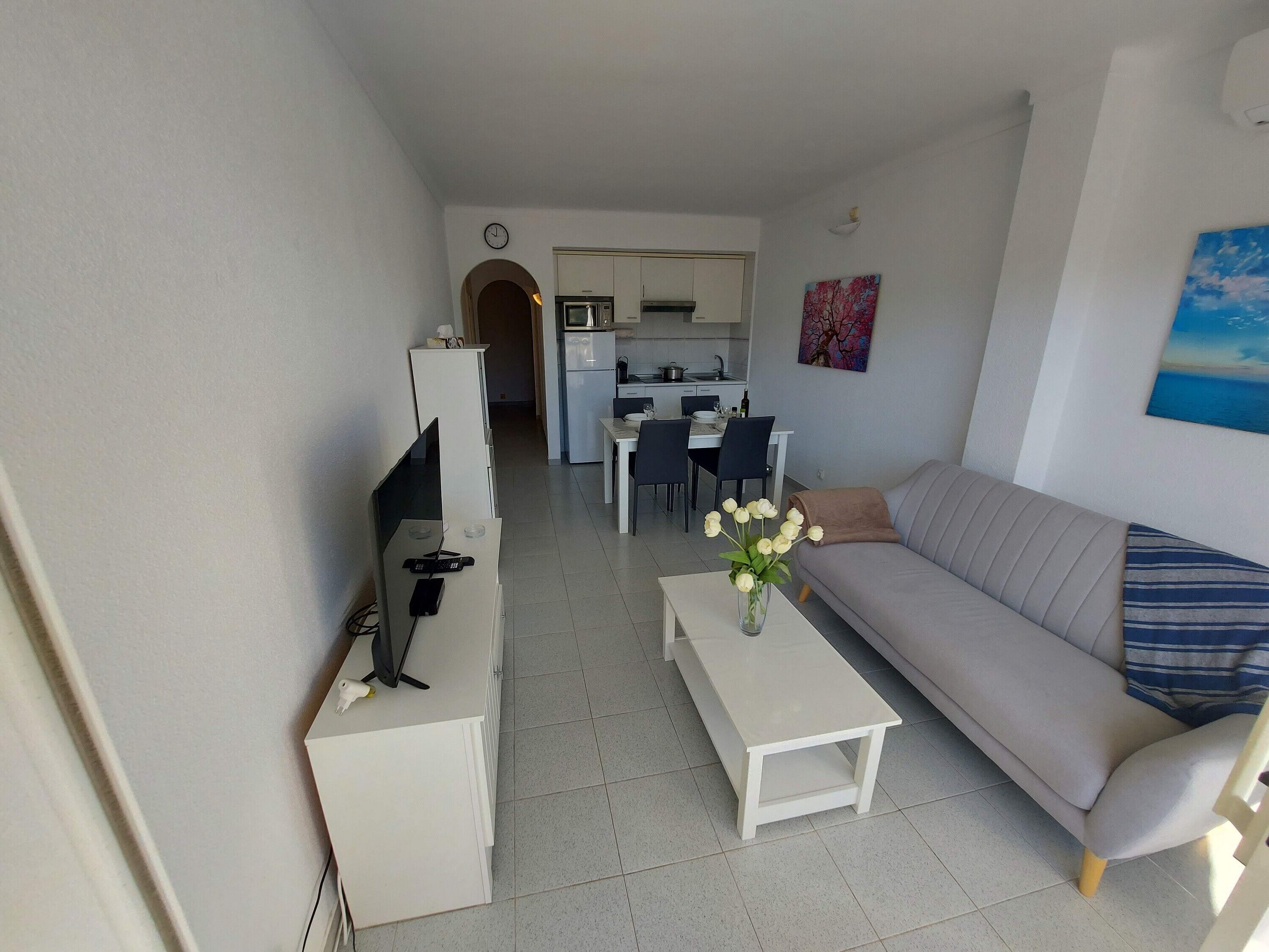 A living room with a sofa, a coffee table, and a TV on a stand. There is a kitchen in the background with a dining table and chairs.