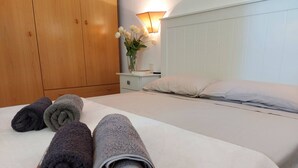 Siististi pedattu sänky, jossa on rullalle taitetut pyyhkeet, yöpöytä lampun kanssa ja puinen vaatekaappi.