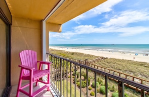 Ein Balkon mit einem pinkfarbenen Adirondack-Stuhl, mit Blick auf einen Strand und eine Holzpromenade.