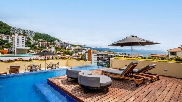 Un área de piscina en la azotea con sillones de descanso y una mesa, rodeada de edificios y vistas panorámicas de la ciudad.