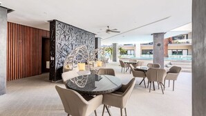 Un espacio moderno al aire libre con comedor, una mesa de vidrio rodeada de sillas, una pared decorativa con patrones geométricos y vistas a una piscina y edificios al fondo.