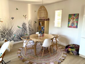 Un coin repas avec une table ronde en bois, des chaises blanches, un tapis tissé et une bibliothèque contenant des livres et des objets décoratifs.
