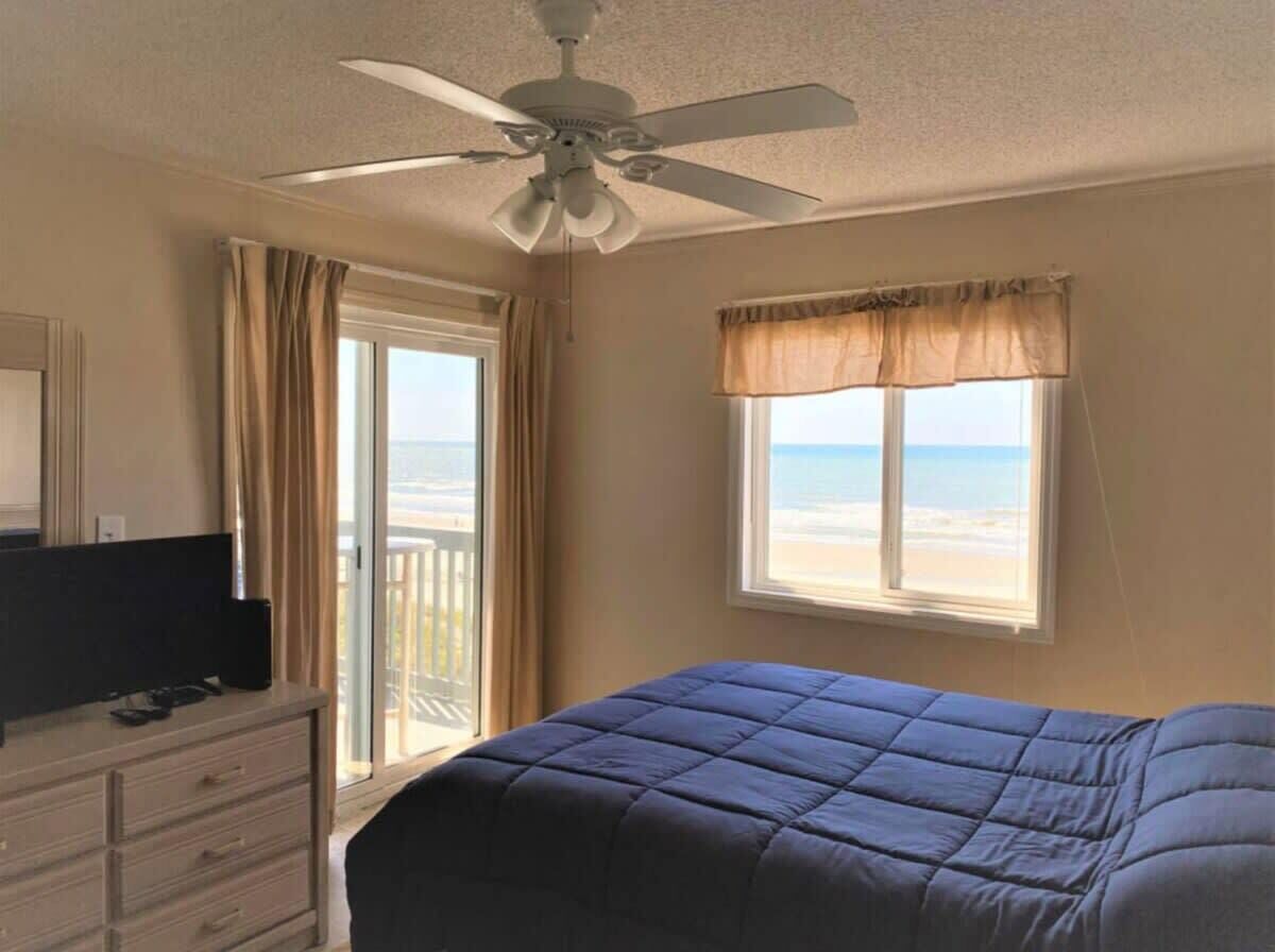 Une chambre à coucher comprenant un lit, un ventilateur de plafond, une commode, un téléviseur et une fenêtre donnant sur la plage.