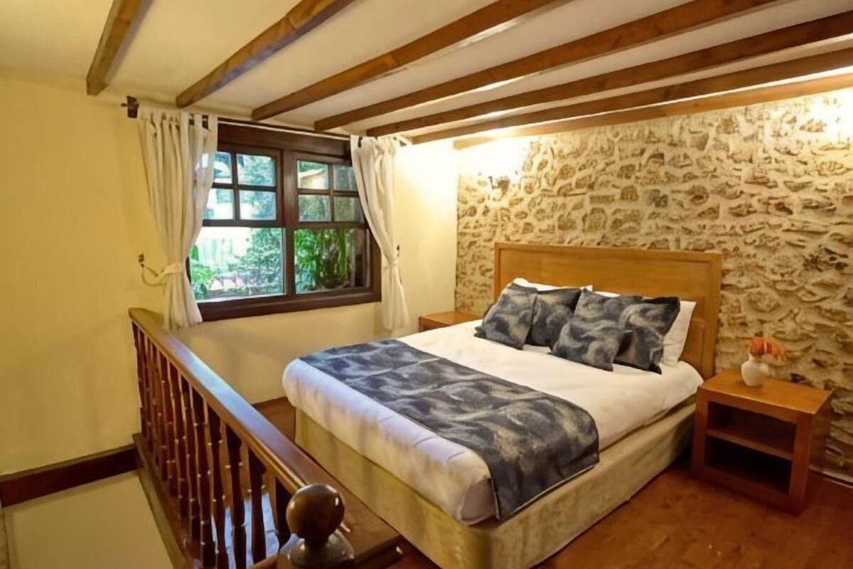 Une chambre avec un mur en pierre, une tête de lit en bois, un lit avec une literie bleue, une table de chevet et une fenêtre avec des rideaux.