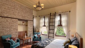 Quarto com parede de tijolos, cama com colcha estampada, poltrona azul, mesa de cabeceira de madeira e um lustre.
