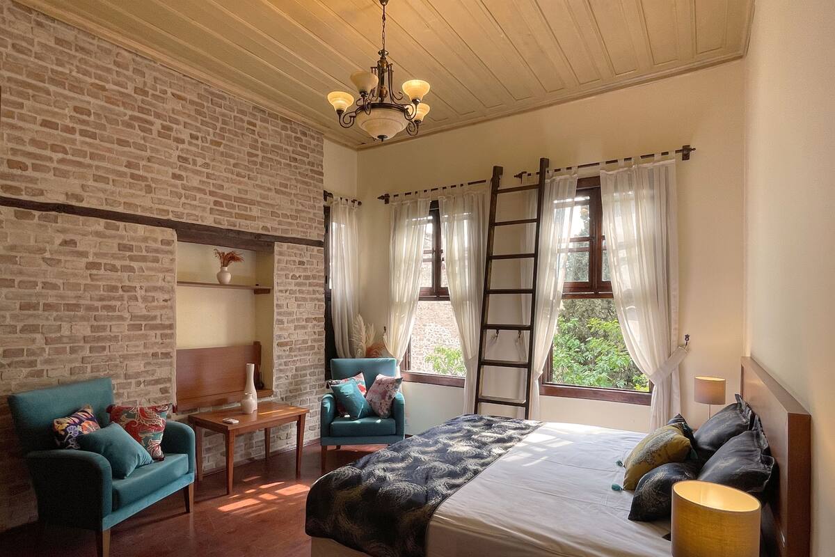 Une chambre avec un mur en briques, un lit recouvert d’une couvre-lit à motifs, un fauteuil bleu, une table de chevet en bois et un lustre.