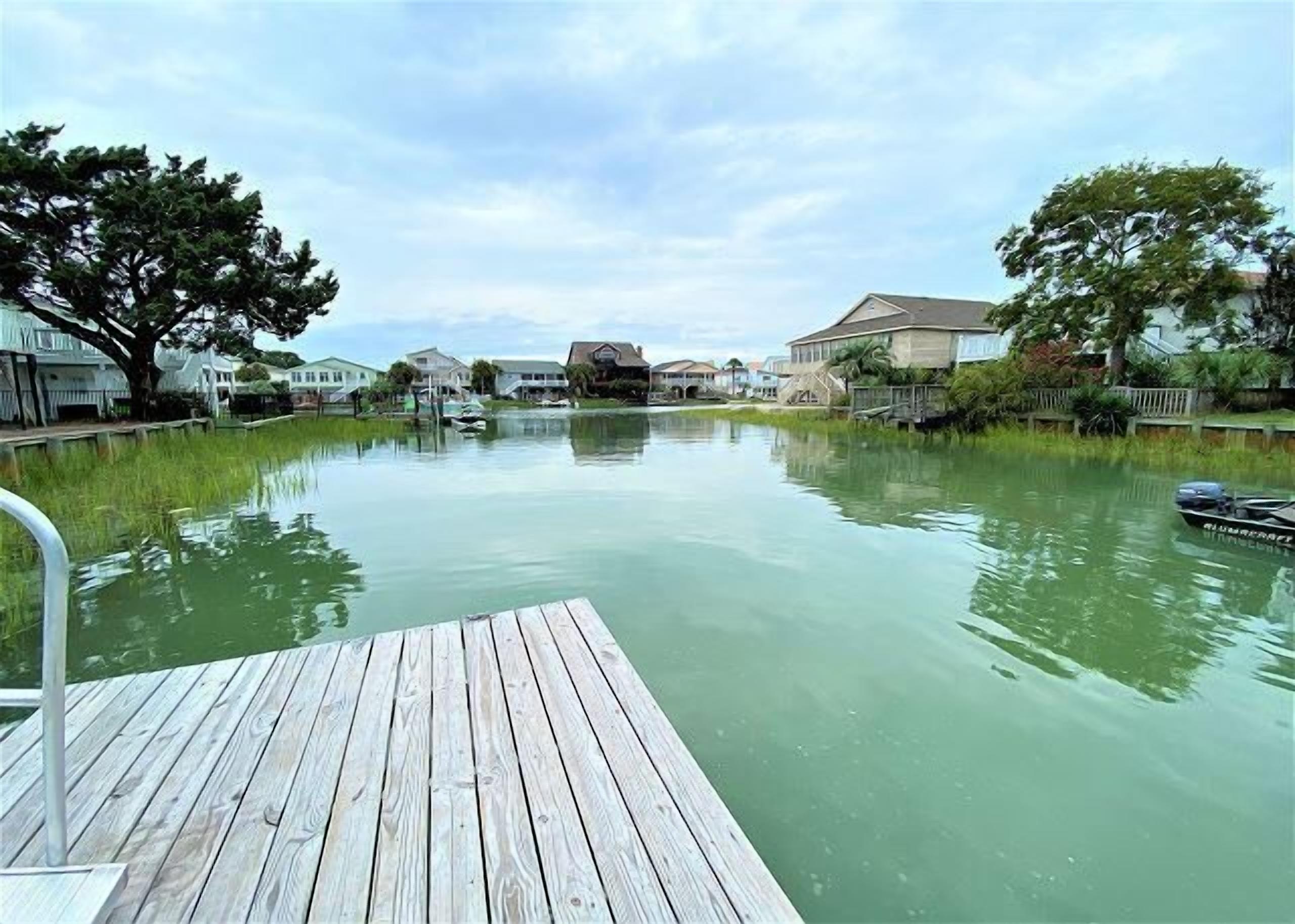 A wooden dock extending over calm water with houses on the opposite bank.