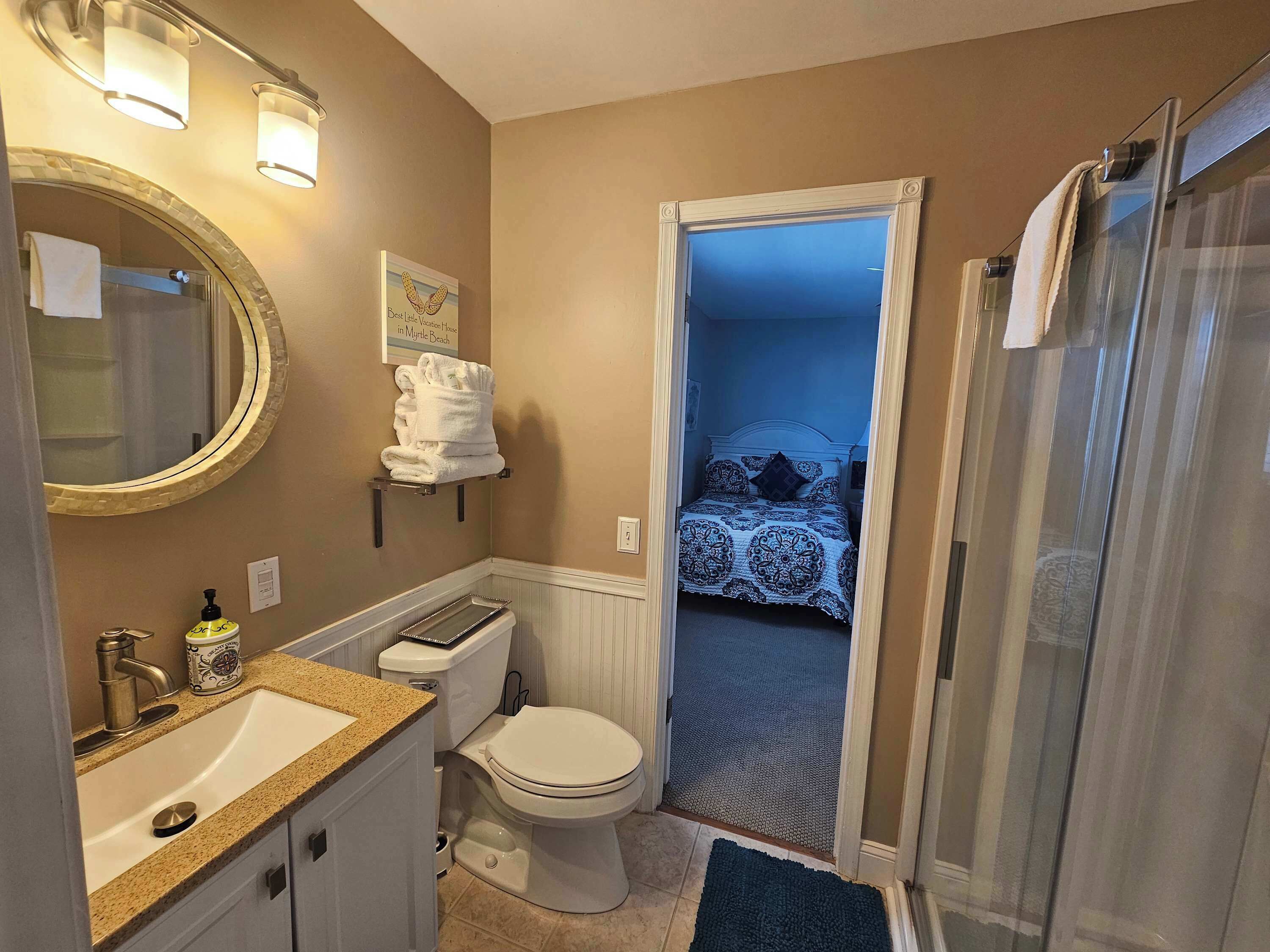 A bathroom with a sink, toilet, and shower. There is a towel rack with towels and a framed picture on the wall.