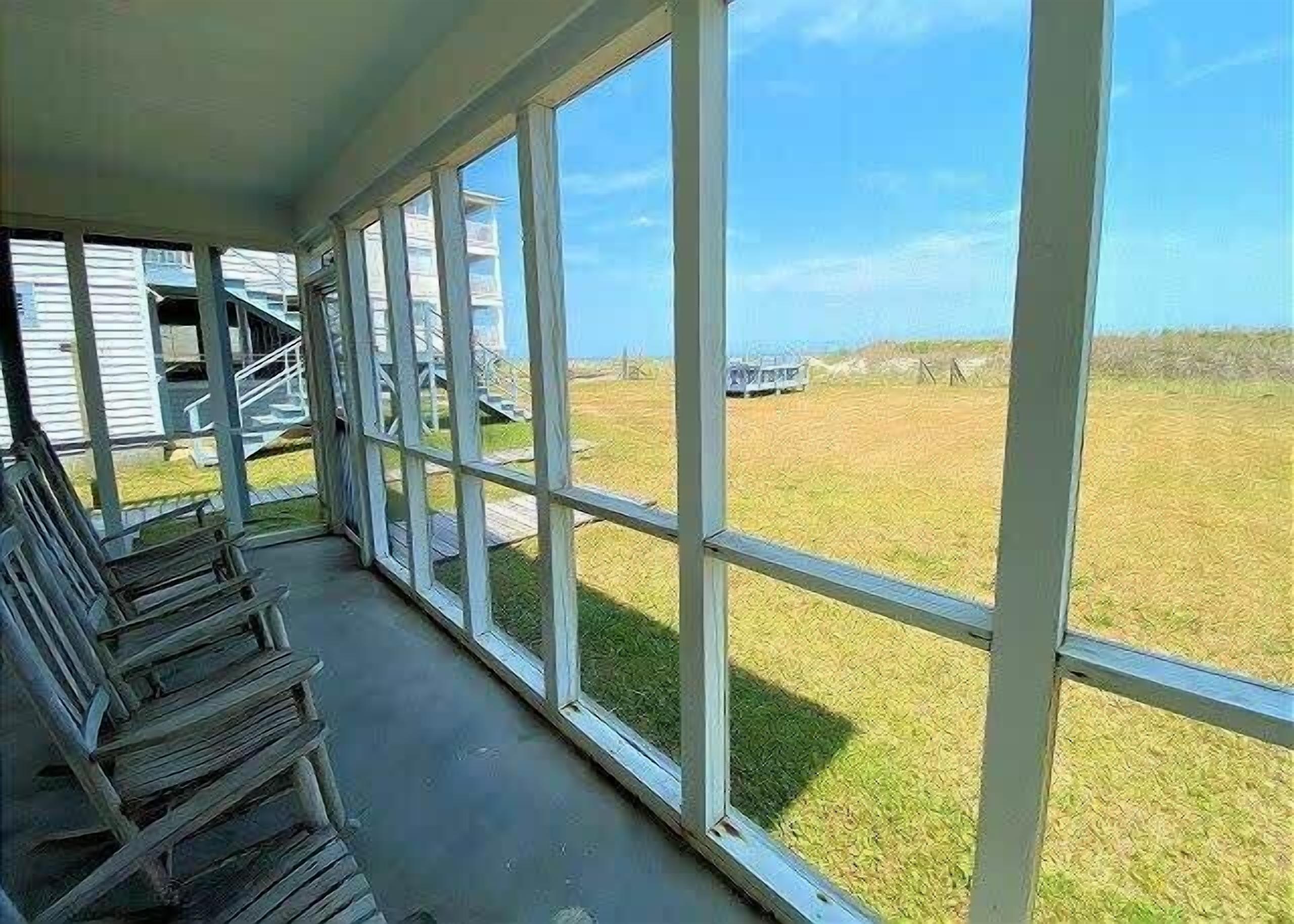 A covered porch with large windows overlooking a grassy field and a building in the distance.
