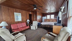 A cozy cabin interior with a wooden ceiling, a red sofa, two armchairs, a dining table, and a ceiling fan. - Cool Air Cottage- Upstairs- Ocean Front Duplex (North Myrtle Beach)