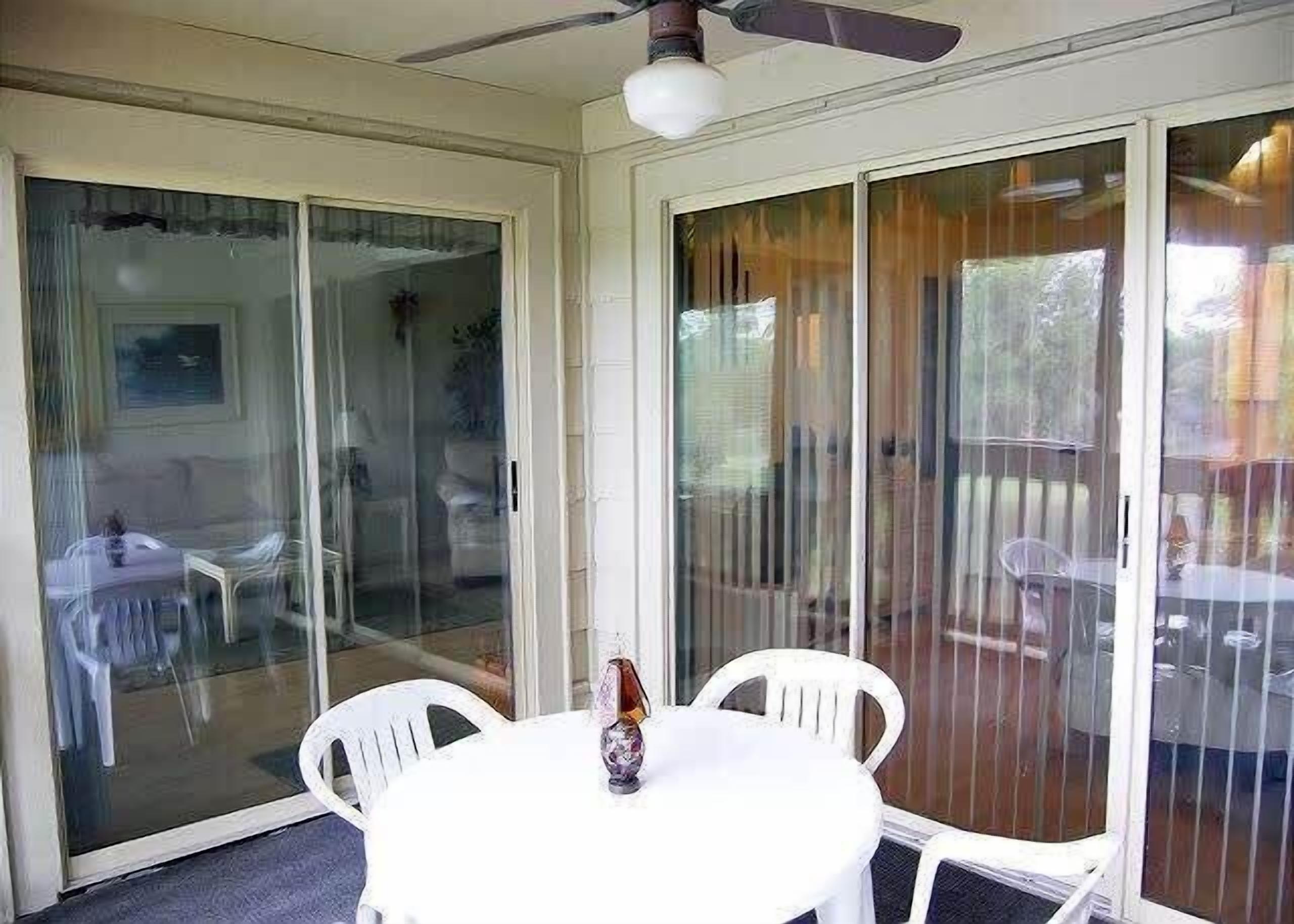 A room with a white table, white chairs, a ceiling fan, and a sliding glass door leading to an outdoor area with a table and chairs.