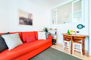 A bright living room with a red sofa, a small wooden table with chairs, a bookshelf, and a window with white frames.
