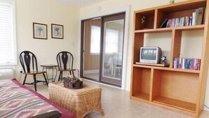 A room with a bed, two chairs, a small table, a wicker basket, a television on a wooden shelf, and a sliding glass door. - It'll Do Two -  Private Pool Close to Beach (North Myrtle Beach)