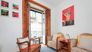A cozy living room with a wooden chair, a patterned sofa, and a small table. A poster is on the wall, and a window with a wooden frame is open. - Cozy Apartment in Historical Lisbon by COZYSTAYS 3/5 (Lisbon)