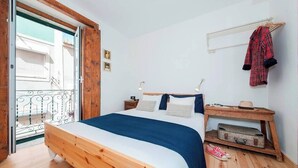 A bedroom with a wooden bed, a nightstand, a suitcase, and a balcony with a railing. - Cozy Apartment in Historical Lisbon by COZYSTAYS 3/5 (Lisbon)