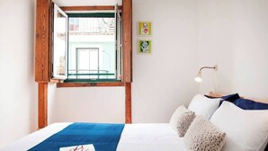 A bedroom with a bed, a window with a wooden frame, a bedside lamp, and two framed pictures on the wall. - Cozy Apartment in Historical Lisbon by COZYSTAYS 4/5 (Lisbon)
