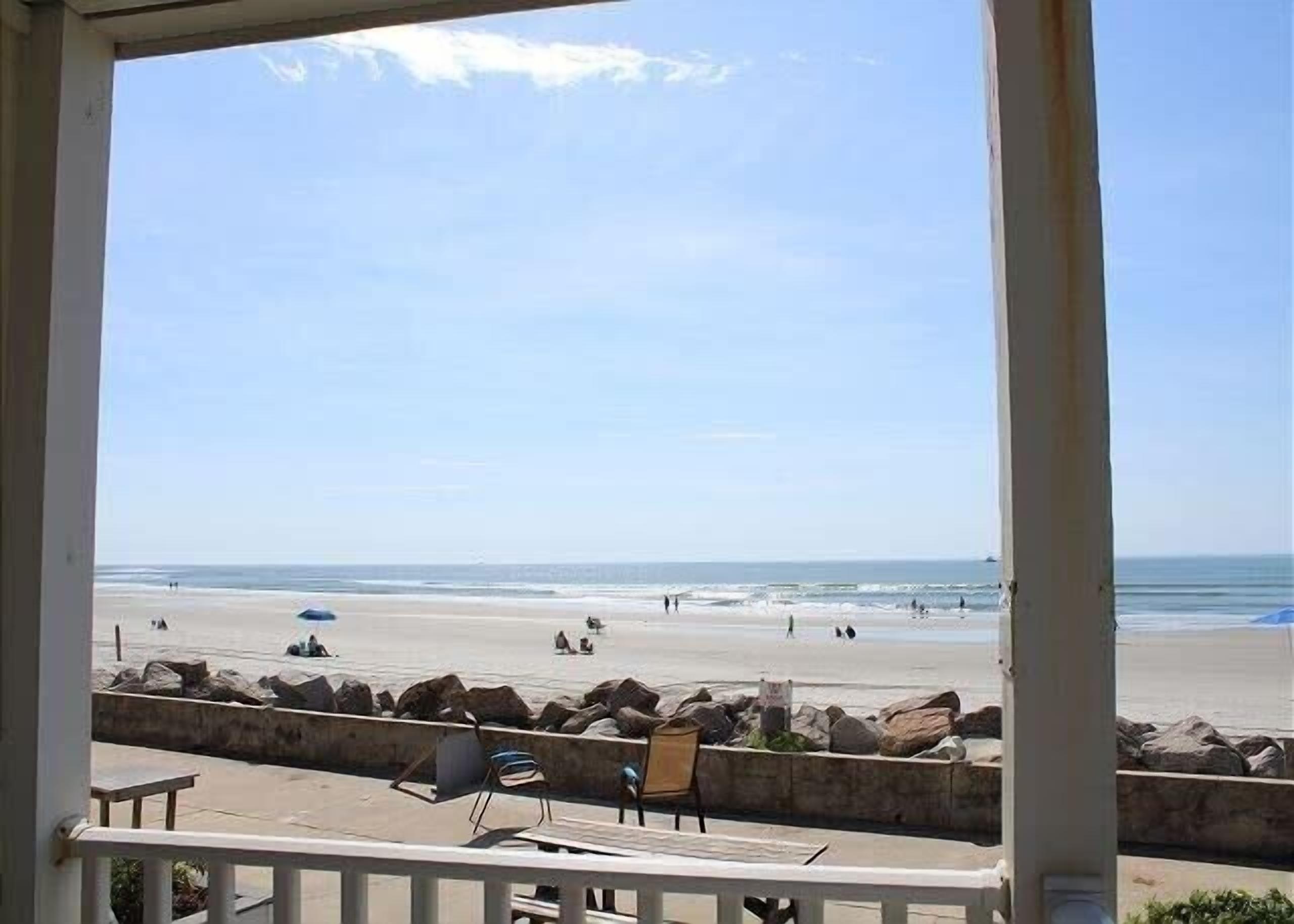 A beach view from a balcony with a railing, a few chairs, and a stone wall.