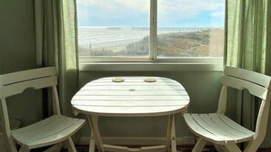 A room with a round table and two white chairs, a window with a view of a bridge and landscape, and green curtains. - Ocean View Villas E2 - The Sea Shed- Oceanview Condo (North Myrtle Beach)
