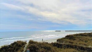 A coastal landscape with a beach, grassy dunes, and a pier extending into the water. - Ocean View Villas E2 - The Sea Shed- Oceanview Condo (North Myrtle Beach)