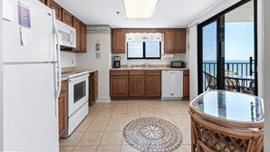 Een compacte keuken met houten kastjes, een witte koelkast, een magnetron en een vaatwasser. Er staat een rond glazen tafel met een rieten stoel, en er is uitzicht op de zee door een schuifdeur van glas.