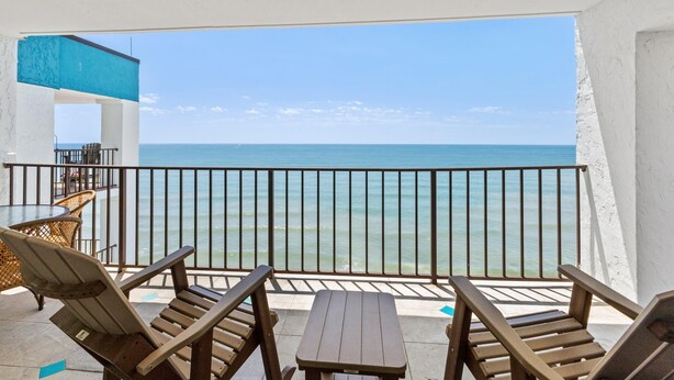 Een balkon met houten stoelen en een tafel, met uitzicht op een strand en een heldere lucht.