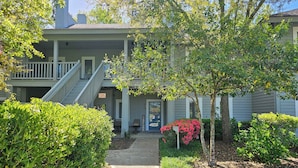 A two-story building with a covered porch, a staircase, and a small garden area with bushes and flowering plants.