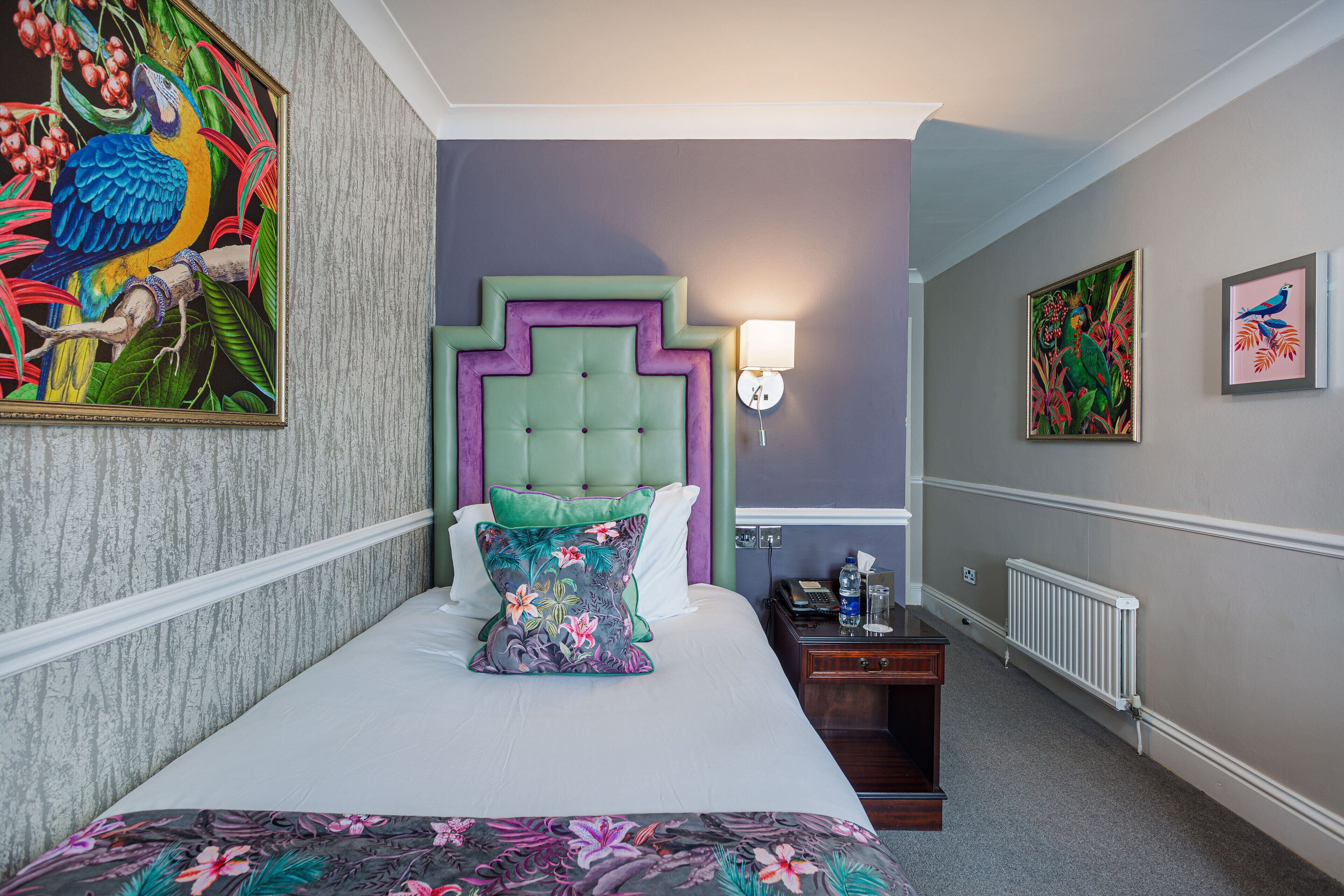 Quarto de hotel com uma cama, mesa de cabeceira, luminária fixada na parede e duas obras de arte emolduradas.
