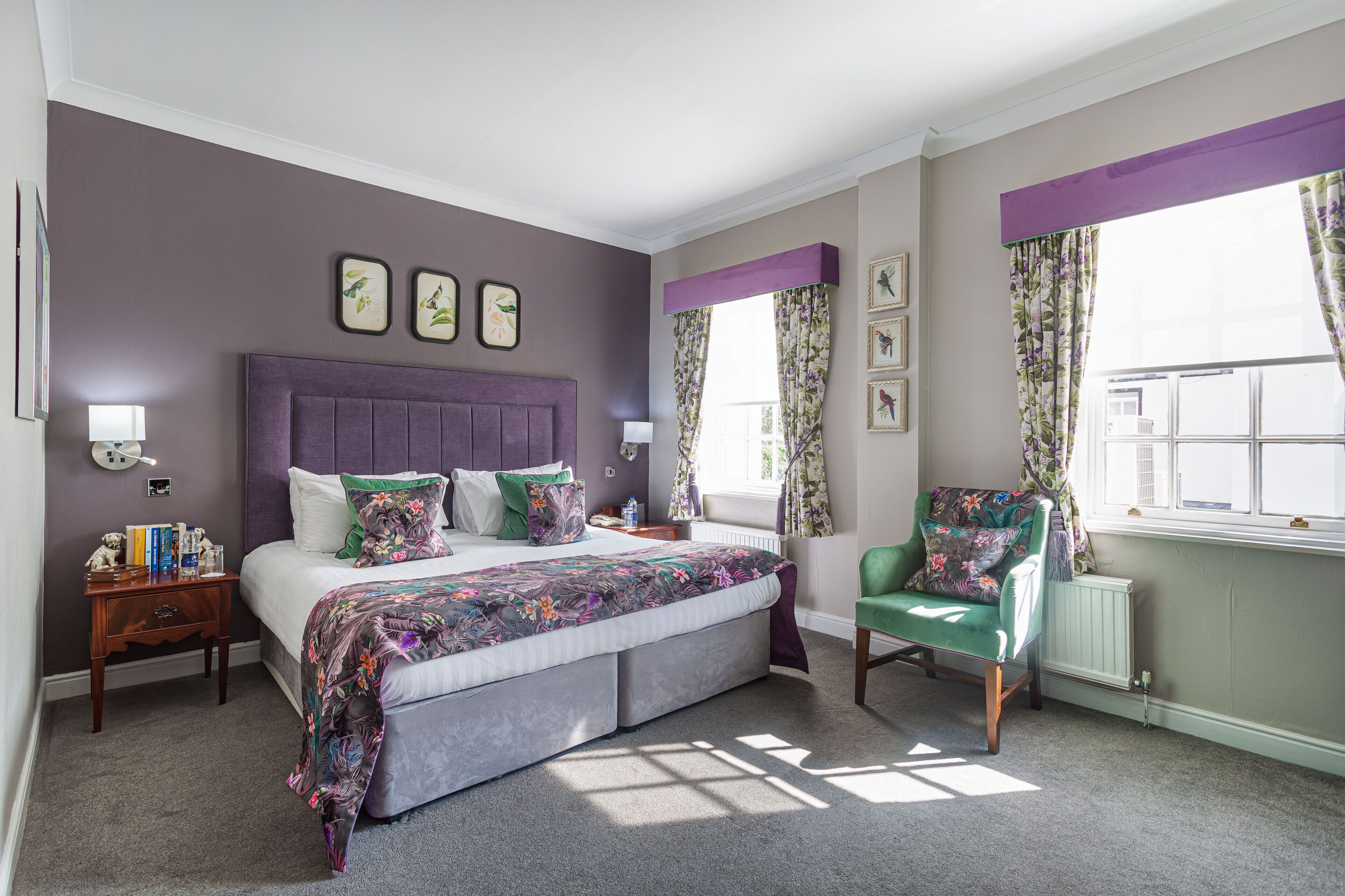 A bedroom with a large bed, a green armchair, a nightstand, and a window with floral curtains.