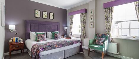 A bedroom with a large bed, a green armchair, a nightstand, and a window with floral curtains.