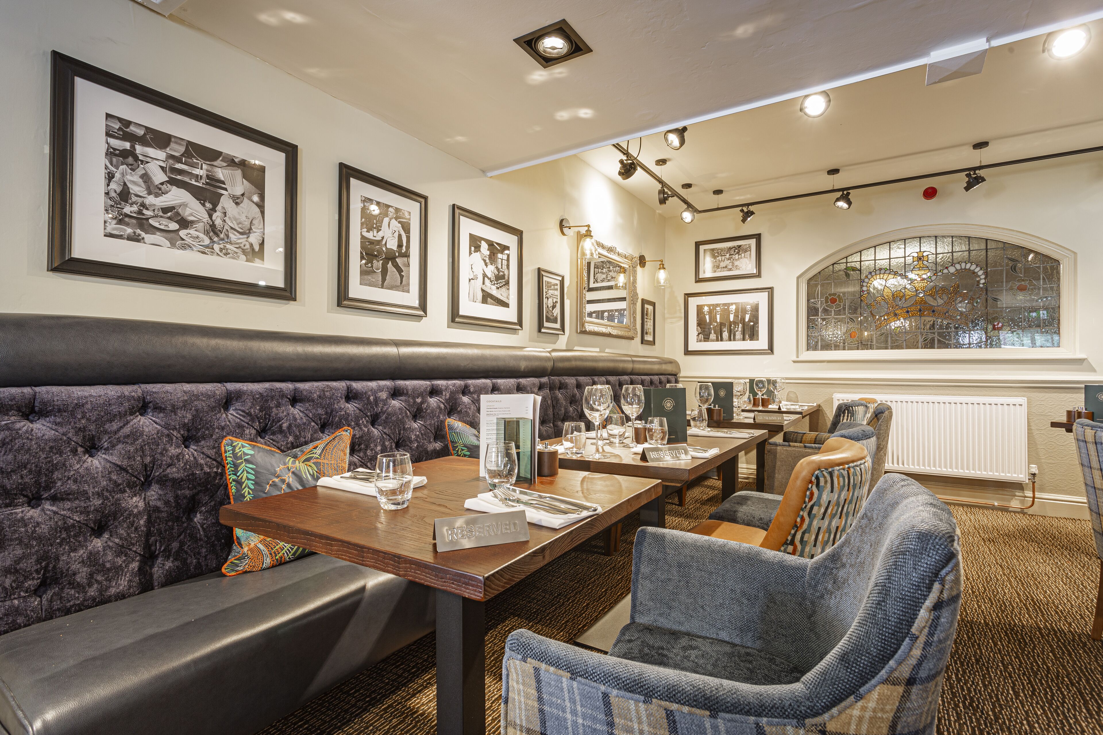 A restaurant with a tufted booth seating area, framed black and white photographs on the walls, and a large arched window with a stained glass design.