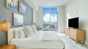 A bedroom with a large bed, a TV, a dresser, and a city view through the window.