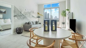 A modern living room with a round dining table, two chairs, a sofa, a large window with a city view, and a wall mural.