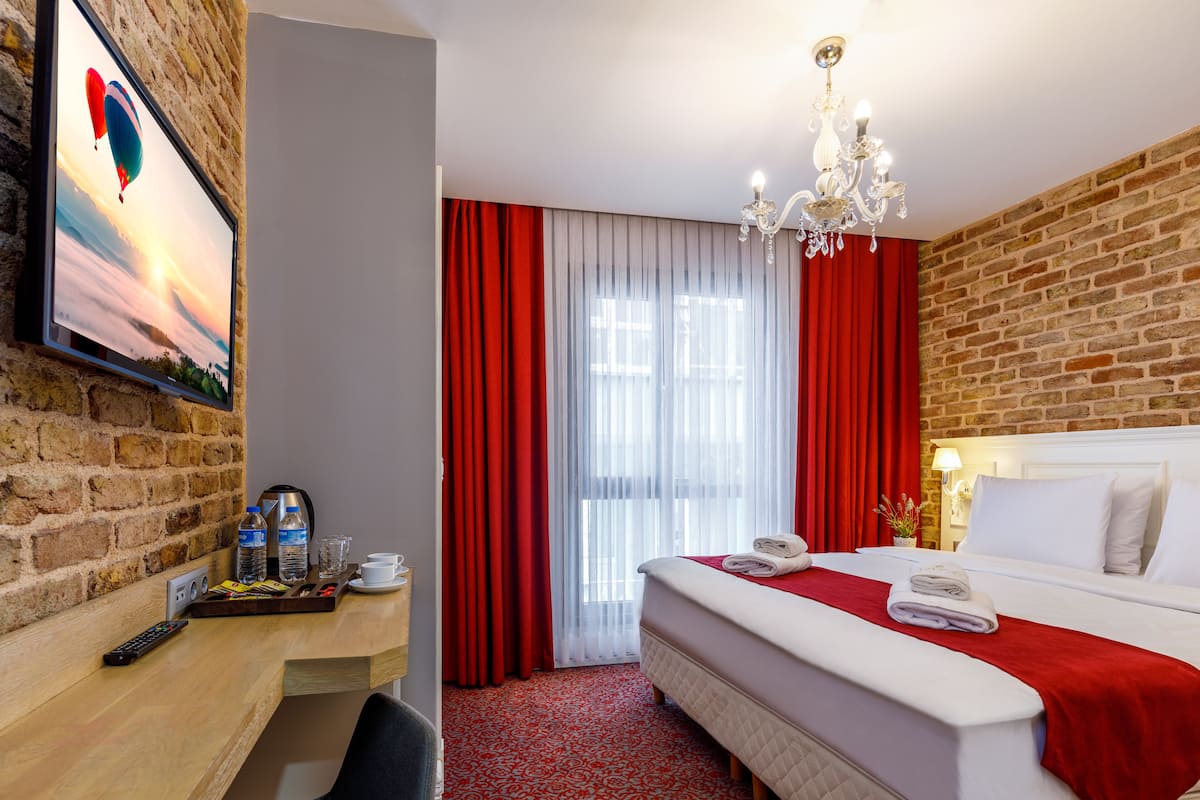 A hotel room with a brick wall, a bed with a red blanket, a chandelier, a desk with a remote control, and a TV displaying a hot air balloon.