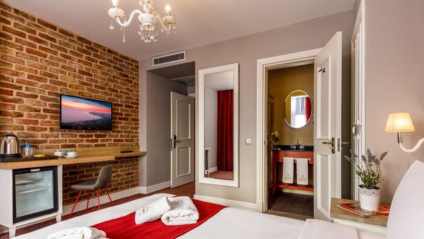 A hotel room with a brick wall, a TV, a desk, a bed with white linens, a red throw, and a bathroom with a mirror and towel rack. - La Porte de Taksim (Istanbul)