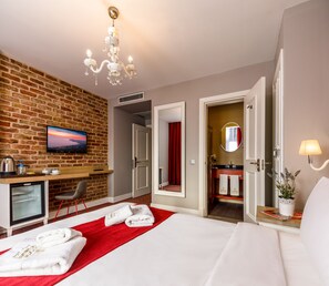 A hotel room with a brick wall, a TV, a desk, a bed with white linens, a red throw, and a bathroom with a mirror and towel rack. - La Porte de Taksim (Istanbul)