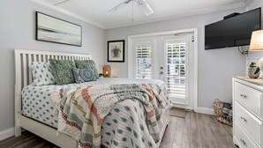 A bedroom with a bed, a ceiling fan, a TV, and a door with shutters.