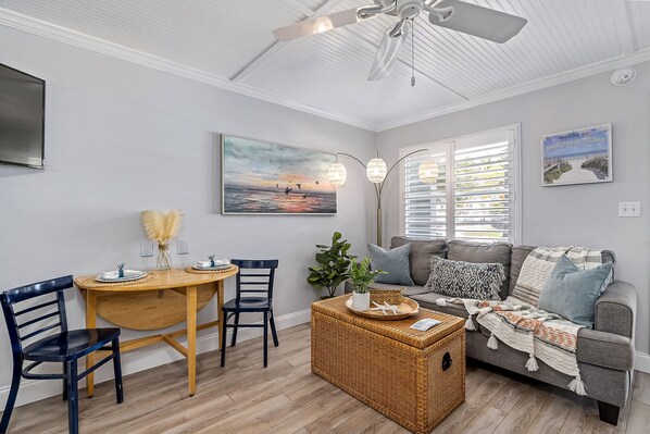 Un salon avec un ventilateur de plafond, une table à manger avec des chaises, un canapé avec des coussins et un tableau accroché au mur.