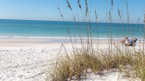 Ein Strand mit Sand und hohem Gras im Vordergrund, und eine Person, die im Hintergrund auf einer Strandmatte liegt.