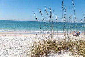 A beach with sand and tall grass in the foreground, and a person lying on a beach towel in the background. - Villas By The Sea #2, Anna Maria Island Condo, one block from beach, heated pool (Bradenton Beach)