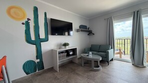 Un salon moderne avec un téléviseur à écran plat, un canapé vert, une petite table ronde et une œuvre d’art représentant un cactus accrochée au mur.