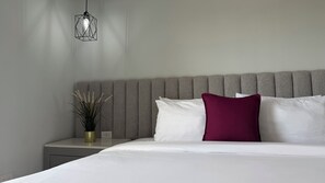 Un lit bien fait, avec des oreillers blancs et bordeaux, une tête de lit rembourrée de couleur grise et un luminaire suspendu.