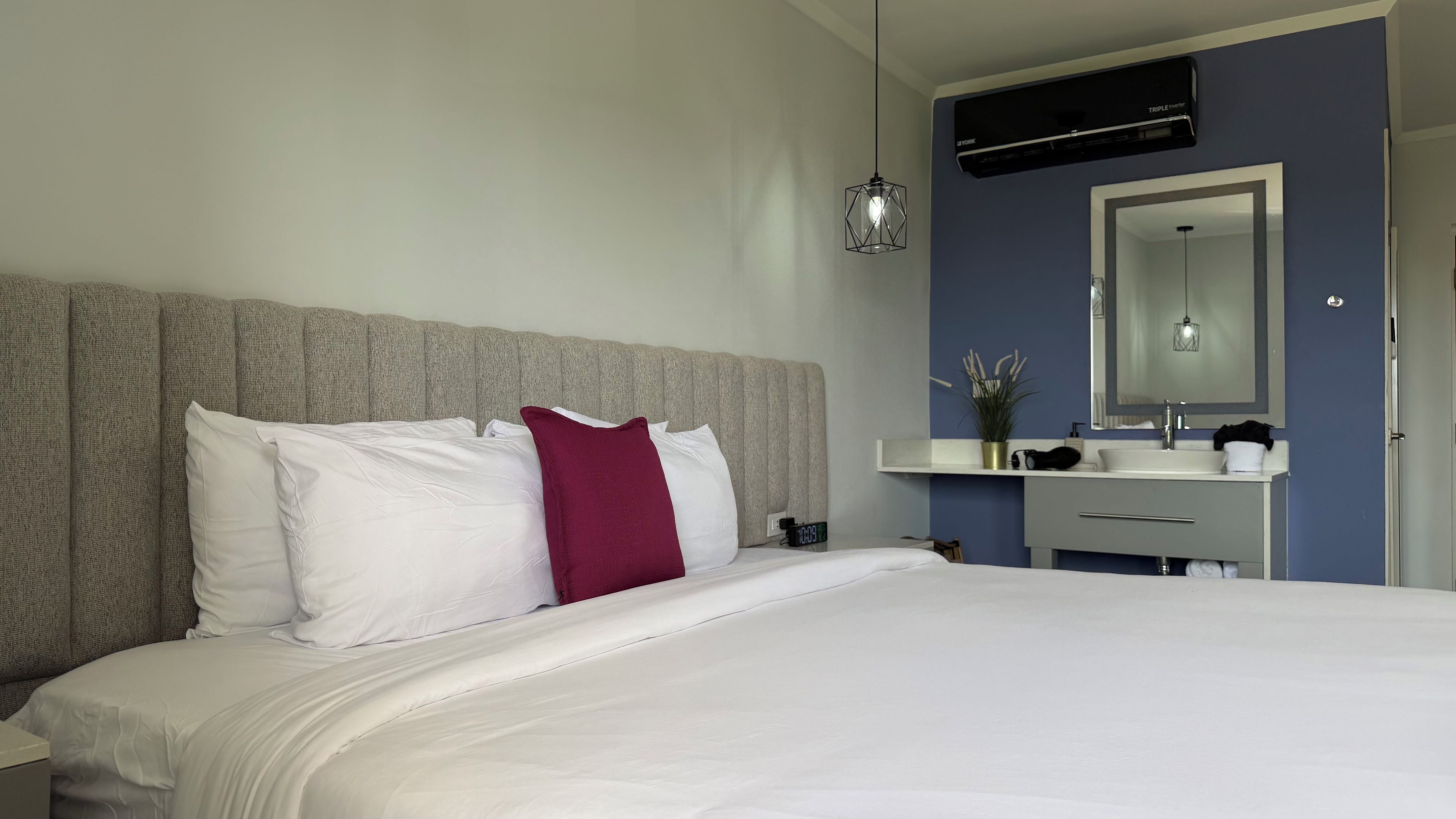 A hotel room with a bed, a bedside table, a nightstand, a wall-mounted air conditioner, a mirror, and a bathroom vanity.