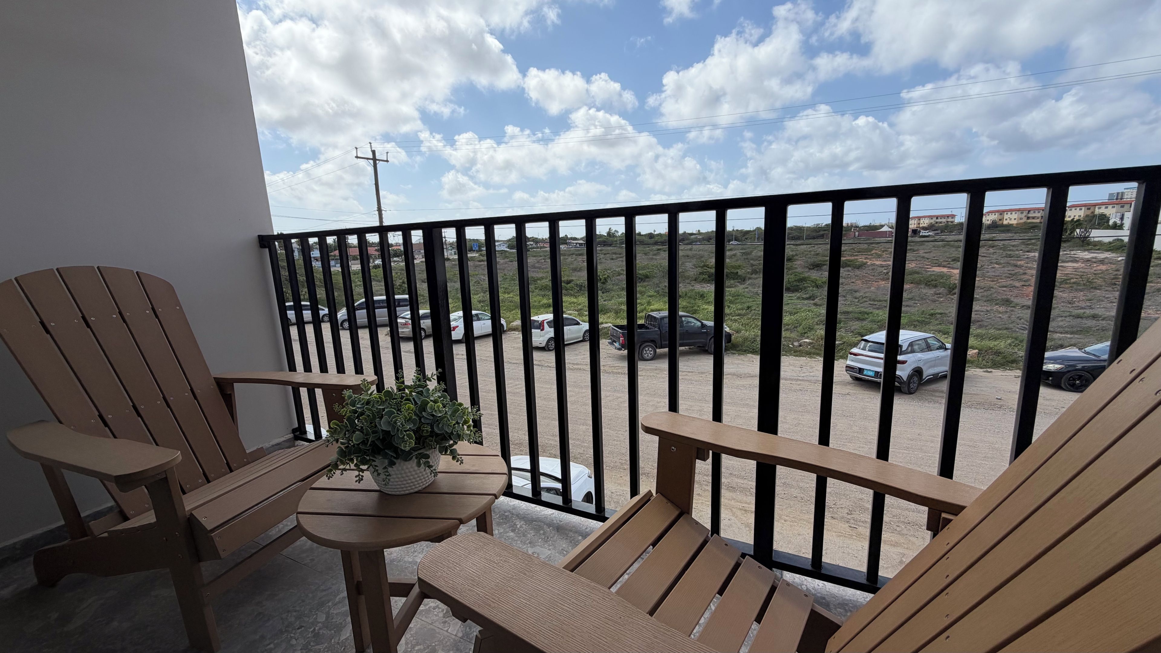 A balcony with wooden chairs and a small table with a plant, overlooking a parking area with several cars.