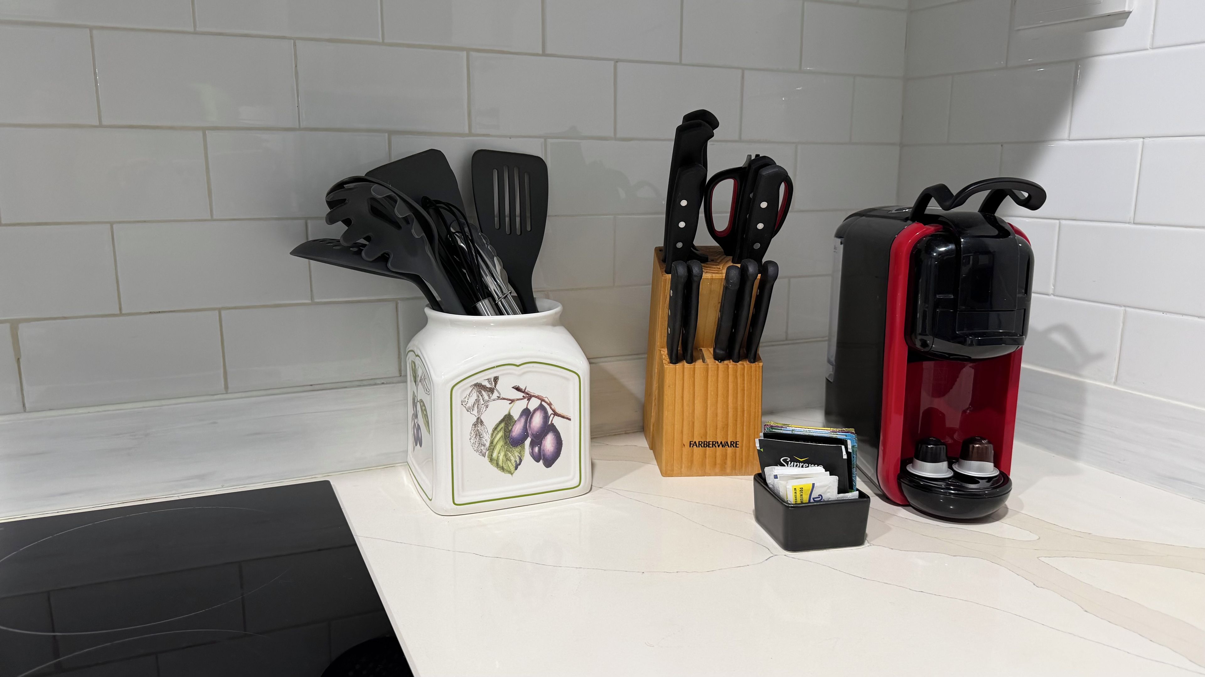 A kitchen counter with a white vase containing kitchen utensils, a wooden knife block, a red coffee maker, and a small box.