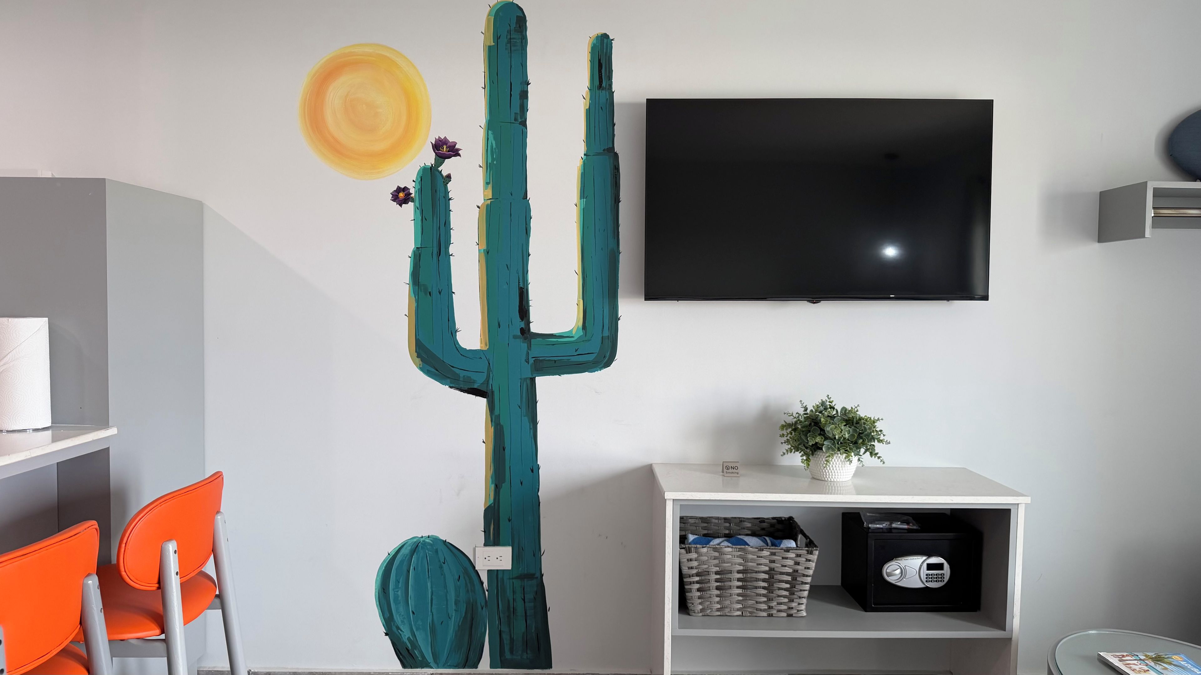 Una habitación con una gran obra de arte de cactus, un televisor de pantalla plana, un estante blanco con una planta y sillas naranjas.
